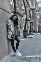 Streetstyle. Volle Länge der schönen jungen Frau in Jeansjacke, die die Hand im Haar hält, während sie sich im Freien an das Gebäude lehnt foto