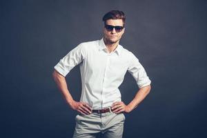 wie Sie selbstbewussten jungen gutaussehenden Mann mit Sonnenbrille machen, der die Arme in die Seite hält und in die Kamera schaut, während er vor grauem Hintergrund steht foto