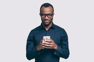 Geschäftsnachricht eingeben. hübscher junger afrikanischer mann, der smartphone hält und es betrachtet, während er vor grauem hintergrund steht foto