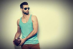 sportlich gutaussehend. Selbstbewusster junger gutaussehender Mann mit Sonnenbrille, der einen American-Football-Ball hält und wegschaut, während er vor weißem Hintergrund steht foto