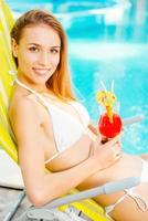 Sommerzeit am Pool verbringen. Seitenansicht einer attraktiven jungen Frau im weißen Bikini, die einen Cocktail trinkt und lächelt, während sie sich im Liegestuhl in der Nähe des Pools entspannt foto