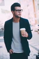 unterwegs Kaffee trinken. hübscher junger Mann in eleganter Freizeitkleidung, der die Straße entlang geht, während er eine Kaffeetasse hält foto