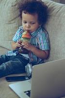 Zeichentrickfilme online anschauen. Blick aus der Vogelperspektive auf den kleinen afrikanischen Jungen, der aus der Babyflasche trinkt und auf seinen Laptop schaut, während er zu Hause auf der Couch sitzt foto