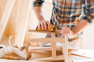 hölzerne Schöpfung. Nahaufnahme eines jungen männlichen Tischlers, der in seiner Werkstatt einen Holzstuhl schleift foto