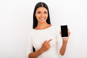 Ich empfehle Ihnen dieses Smartphone. lächelnde junge Frau, die Handy hält und darauf zeigt, während sie vor weißem Hintergrund steht foto