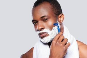 Mann rasiert. Schöner hemdloser afrikanischer Mann, der sein Gesicht rasiert und in die Kamera schaut, während er vor grauem Hintergrund steht foto