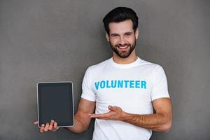 Wohltätigkeit ist einfacher mit Technologien. Selbstbewusster junger Mann im Freiwilligen-T-Shirt, der sein digitales Tablet zeigt und lächelnd in die Kamera blickt, während er vor grauem Hintergrund steht foto