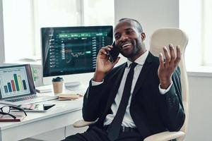 glücklicher junger afrikanischer mann in formalwear, der am telefon spricht und lächelt, während er im büro arbeitet foto