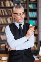 Zeit für eine kleine Pause nehmen. Selbstbewusster älterer Mann mit grauen Haaren in formeller Kleidung, der eine Kaffeetasse hält und wegschaut, während er sich an den Tisch lehnt und mit einem Bücherregal im Hintergrund foto