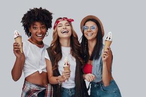 Drei attraktive junge Frauen essen Eis und lächeln im Stehen vor grauem Hintergrund foto