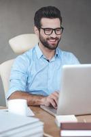 tolle lösung. Nahaufnahme fröhlicher junger gutaussehender Mann mit Brille, der am Laptop arbeitet und lächelt, während er an seinem Arbeitsplatz sitzt foto
