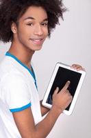 Arbeiten an einem digitalen Tablet. Rückansicht eines fröhlichen afrikanischen Teenagers, der an einem digitalen Tablet arbeitet und über die Schulter schaut, während er isoliert auf grauem Hintergrund steht foto