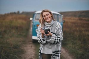 junger Fotograf. Attraktive junge Frau, die Digitalkamera hält und lächelt, während sie im Freien mit dem blauen Minivan im Retro-Stil im Hintergrund steht foto