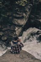 sein Lieblingshobby. Draufsicht des jungen modernen Mannes, der fotografiert, während er auf dem Felsen mit dem Fluss unten sitzt foto