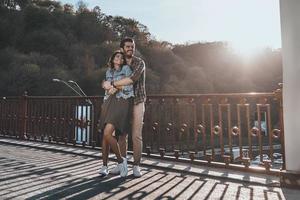 in voller Länge von einem gutaussehenden Mann, der junge attraktive Frau umarmt, während er draußen auf der Brücke steht foto