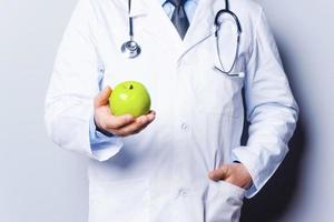 Früchte machen gesund. Nahaufnahme eines Arztes, der grünen Apfel hält, während er vor grauem Hintergrund steht foto