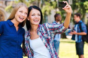 einen glücklichen Moment einfangen. Zwei schöne junge Frauen machen Selfie, während sie nahe beieinander stehen, während zwei Männer im Hintergrund sprechen foto