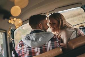 sie ist das Zentrum seines Universums. Schönes junges Paar lächelt, während es von Angesicht zu Angesicht auf den Beifahrersitzen im Minivan im Retro-Stil sitzt foto
