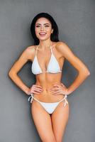 fit und schön. Attraktive junge Frau im weißen Bikini, die Hände auf der Hüfte hält und lächelt, während sie vor grauem Hintergrund steht foto