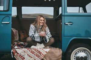schönen Tag genießen. Attraktive junge lächelnde Frau, die einen Becher hält und wegschaut, während sie im Inneren des blauen Minivans im Retro-Stil sitzt foto