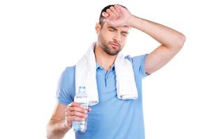 fühle mich so müde. müder junger Mann mit Handtuch auf den Schultern, der eine Flasche mit Wasser hält und lächelt, während er vor weißem Hintergrund steht foto