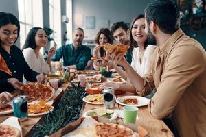 Urlaub unter Freunden. Gruppe junger Leute in Freizeitkleidung, die Pizza essen und lächeln, während sie drinnen eine Dinnerparty haben foto