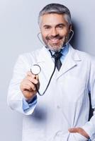 medizinische Untersuchung. Fröhlicher, reifer Arzt mit grauem Haar, der Sie mit Stethoskop untersucht und lächelt, während er vor grauem Hintergrund steht foto