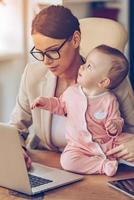 Vollzeit berufstätige Mutter. Kleines Mädchen, das aufschaut, während es mit seiner Mutter im Büro auf dem Schreibtisch sitzt foto