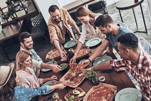 Lieblingsessen. Draufsicht auf junge Leute in Freizeitkleidung, die Pizza pflücken und lächeln, während sie eine Dinnerparty haben foto