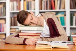 in der Bibliothek schlafen. Müder junger Mann, der seinen Kopf auf dem Bücherstapel hält und schläft, während er am Bibliothekstisch sitzt foto