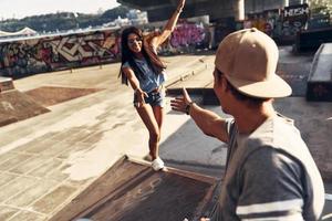 Schöne junge Frau, die versucht, zu ihrer Freundin aufzusteigen, während sie Zeit im Skatepark im Freien verbringt foto