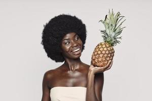 Porträt einer schönen jungen Afrikanerin, die Ananas hält und lächelt, während sie vor grauem Hintergrund steht foto