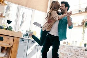 Es ist so toll, in voller Länge mit einem schönen jungen Paar in Freizeitkleidung zusammen zu sein, das tanzt und lächelt, während es zu Hause in der Küche steht foto