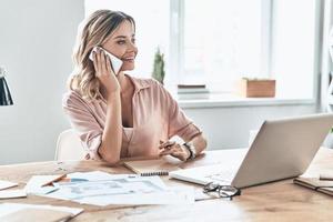 Ich kann es total zum Laufen bringen. Schöne junge Frau in eleganter Freizeitkleidung, die lächelt und telefoniert, während sie in einem modernen Büro sitzt foto