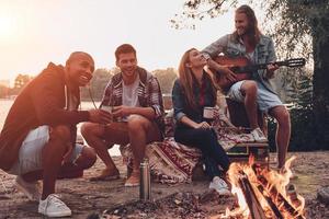 sorglos campen. Gruppe junger Leute in Freizeitkleidung, die lächeln, während sie eine Strandparty am Lagerfeuer genießen foto