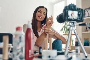 atemberaubende junge Frau, die Lipgloss aufträgt und lächelt, während sie ein Social-Media-Video macht foto