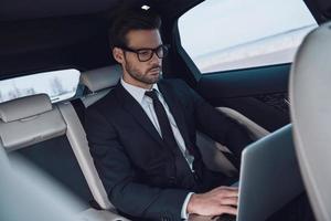 jung und erfolgreich. hübscher junger Mann im vollen Anzug, der mit Laptop arbeitet, während er im Auto sitzt foto