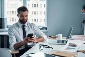 hübscher junger Mann in Hemd und Krawatte mit Smartphone und lächelnd, während er im Büro sitzt foto