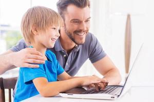 wenig es Genie. Glücklicher Vater und Sohn, die auf den Laptop schauen, während der kleine Junge etwas auf der Tastatur tippt und lächelt foto