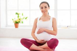 Ruhe bewahren für ihr ungeborenes Baby. Schöne schwangere Frau, die im Lotussitz auf der Trainingsmatte sitzt und lächelt foto