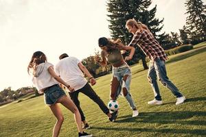 beste Art, das Wochenende zu verbringen. Gruppe junger lächelnder Menschen in Freizeitkleidung, die beim Fußballspielen im Freien um den Ball kämpfen foto