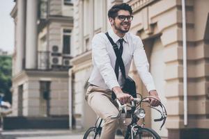 genießt seine Fahrt zur Arbeit. Tiefwinkelansicht eines fröhlichen jungen Mannes mit Brille, der mit einem Lächeln wegschaut, während er mit dem Fahrrad fährt foto