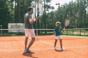 gemeinsam trainieren. Rückansicht des Vaters in voller Länge in Sportkleidung, die seiner Tochter das Tennisspielen beibringt, während beide auf dem Tennisplatz stehen foto
