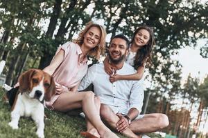 immer übereinander tragen. glückliche junge dreiköpfige Familie mit lächelndem Hund beim Sitzen auf Gras im Park foto