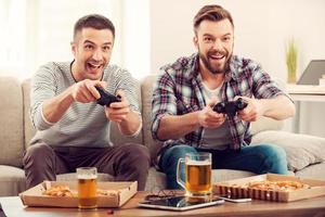 die begeisterten Gamer. Zwei junge glückliche Männer spielen Videospiele, während sie auf dem Sofa sitzen foto