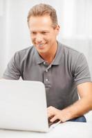 Mann surft im Internet. fröhlicher Mann mit grauen Haaren im Poloshirt mit Laptop und lächelnd foto