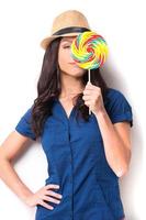 Schönheit mit Süßigkeiten. Verspielte junge schöne Frau mit flippigem Hut, die Lutscherbonbons vor ihrem Auge hält und lächelt, während sie vor weißem Hintergrund steht foto