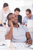 kreative Menschen bei der Arbeit. hübscher junger afrikanischer mann, der kamera hält und lächelt, während drei leute im hintergrund arbeiten foto