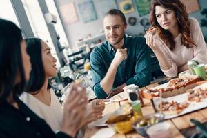 einfach nur Entspannen. Gruppe junger Leute in Freizeitkleidung, die reden und lächeln, während sie drinnen eine Dinnerparty haben foto