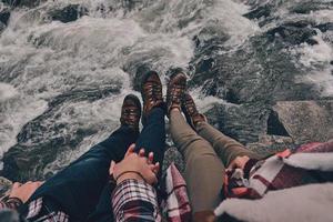 immer zusammen. Nahaufnahme von oben auf ein junges Paar, das Händchen hält, während es auf den Felsen mit dem Fluss darunter sitzt foto